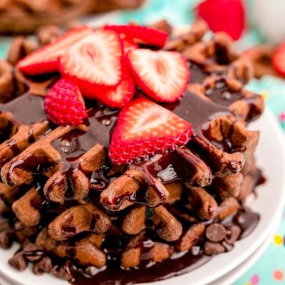 Close up photo of a stack of chocolate waffles with chocolate syrup and sliced strawberries on top.