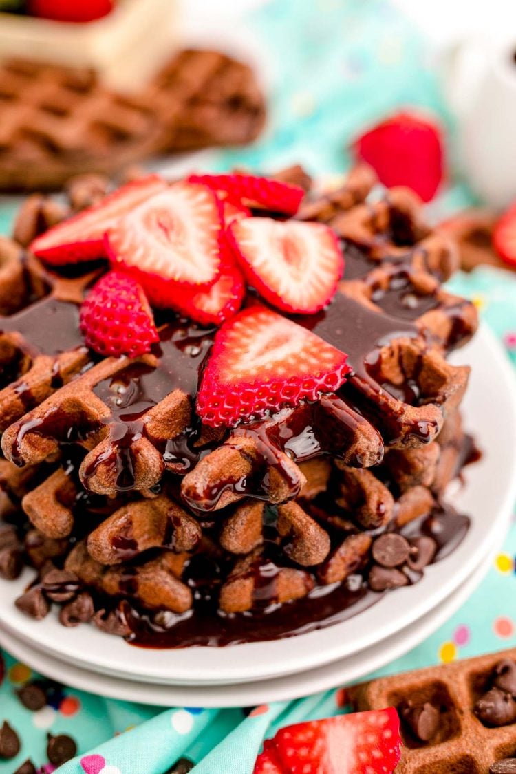 Close up photo of a stack of chocolate waffles with chocolate syrup and sliced strawberries on top.