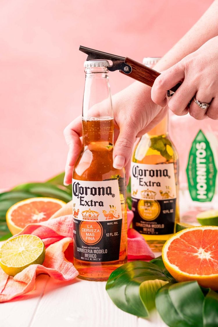 A woman popping the cap of a Corona off with a bottle opener.