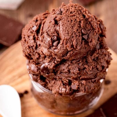 Close up photo of edible brownie batter in a glass bowl on a wooden board with a spoon next to it.