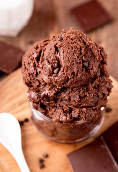 Close up photo of edible brownie batter in a glass bowl on a wooden board with a spoon next to it.