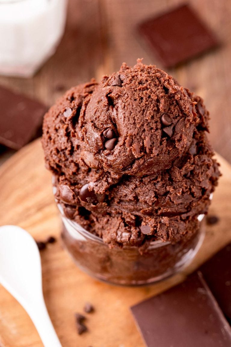 Close up photo of edible brownie batter in a glass bowl on a wooden board with a spoon next to it.