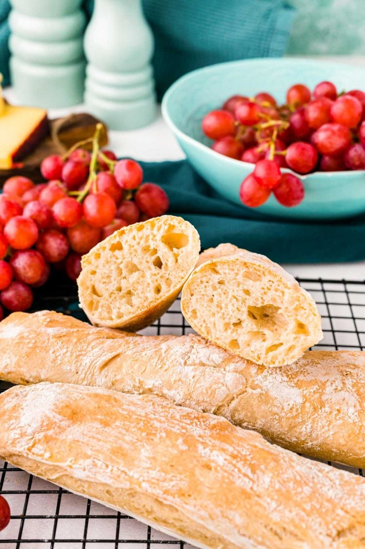 Three French baguettes on a black wire rack, one has been cut in half and you can see the inside. Grapes in the background.