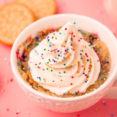 A white tea cup with a golden oreo mug cake topped with buttercream and sprinkles in it.