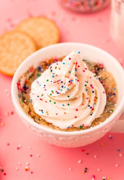 A white tea cup with a golden oreo mug cake topped with buttercream and sprinkles in it.