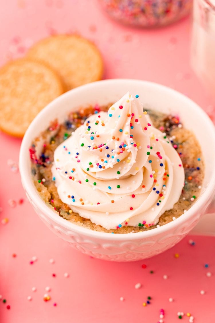 A white tea cup with a golden oreo mug cake topped with buttercream and sprinkles in it.