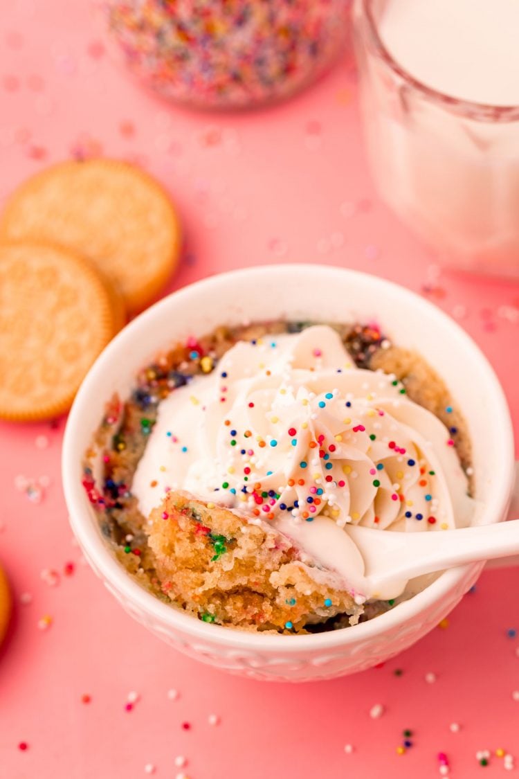 A spoon taking a bite out of a golden Oreo mug cake in a white cup on a pink surface.
