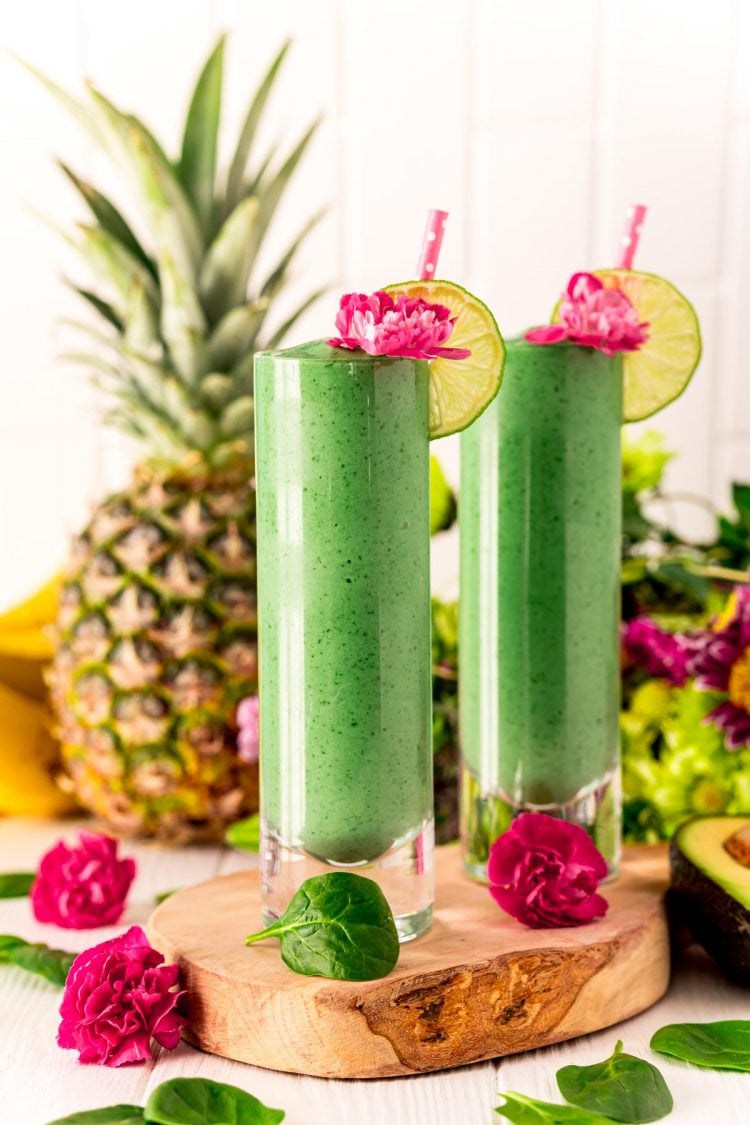 Two green smoothies on a wooden cutting board garnished with pink flowers and limes.