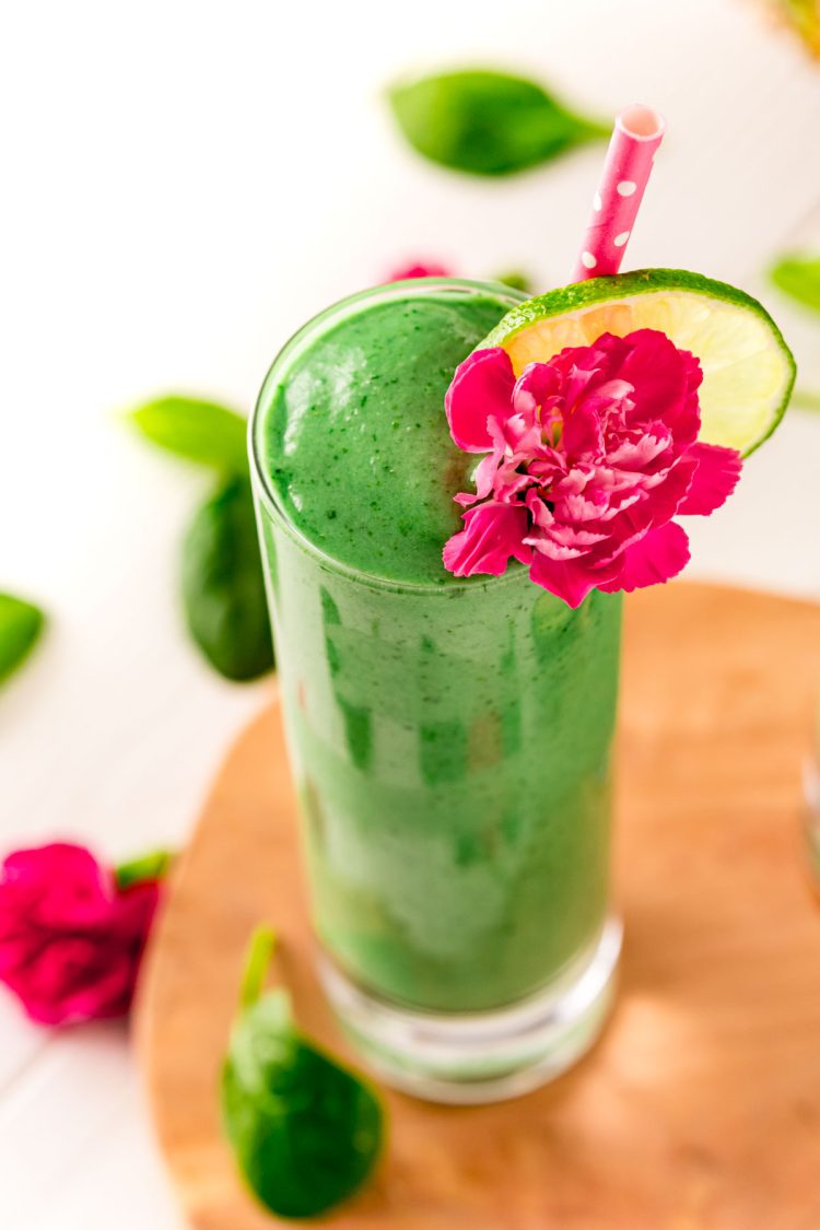 Close up photo of a green smoothie in a tall glass garnished with a pink carnation and lime slice.