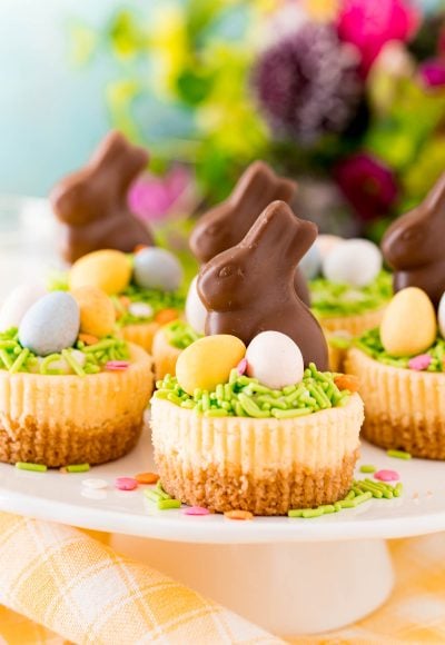 Easter cheesecakes topped with cadbury eggs and chocolate bunnies on a white cake stand.