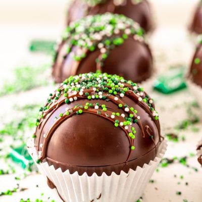 Close up photo of mint hot chocolate bombs on a white surface with green sprinkles and chocolate mints scattered around.