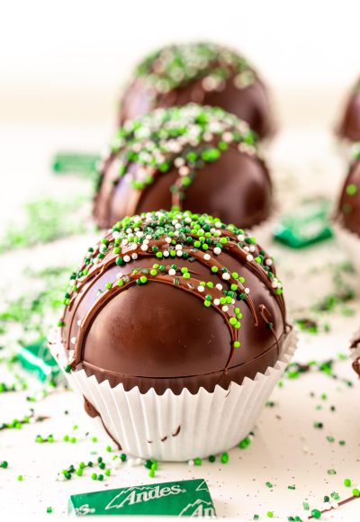 Close up photo of mint hot chocolate bombs on a white surface with green sprinkles and chocolate mints scattered around.