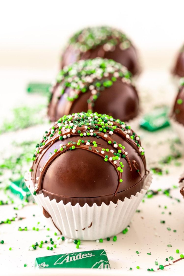 Close up photo of mint hot chocolate bombs on a white surface with green sprinkles and chocolate mints scattered around.