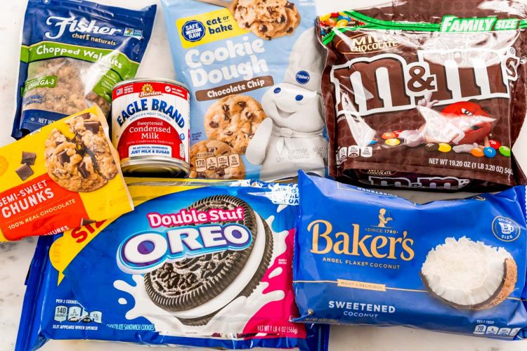 Packages of walnuts, sweetened condensed milk, Oreos, chocolate chunks, coconut, M&Ms, and cookie dough on a white table.