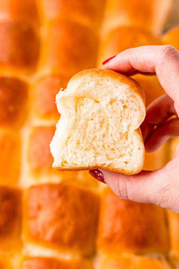 A woman's hand holding up a dinner roll to the camera with more rolls in the background.