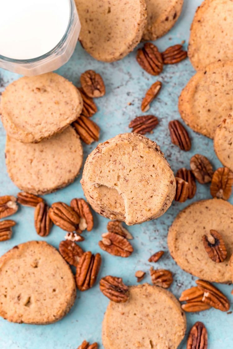 Overhead photo of pecan sandies scattered on a blue surface with pecans and a glass of milk.