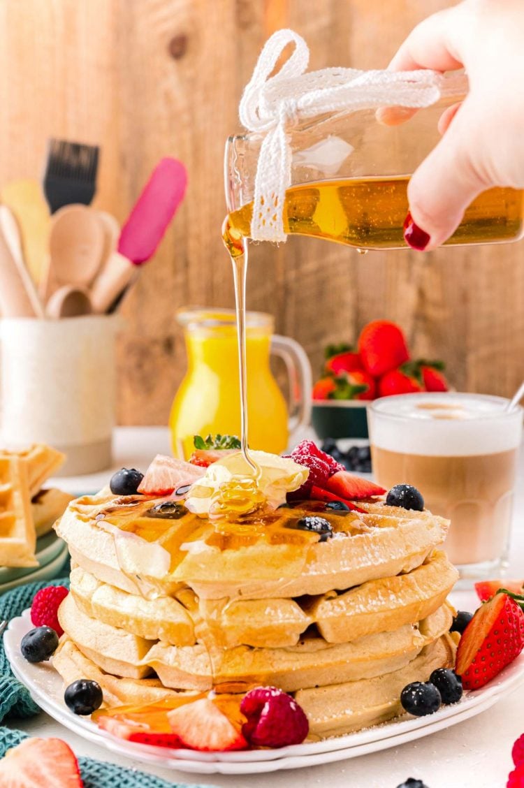 Close up photo of a woman pouring maple syrup over the top of a stack of Belgian waffles.