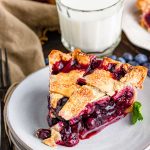 Close up photo of a slice of blueberry pie with a lattice crust on a white plate with more pie and a glass of milk in the background.