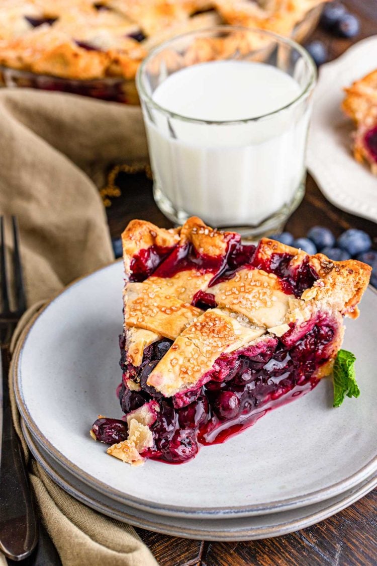 Close up photo of a slice of blueberry pie with a lattice crust on a white plate with more pie and a glass of milk in the background.