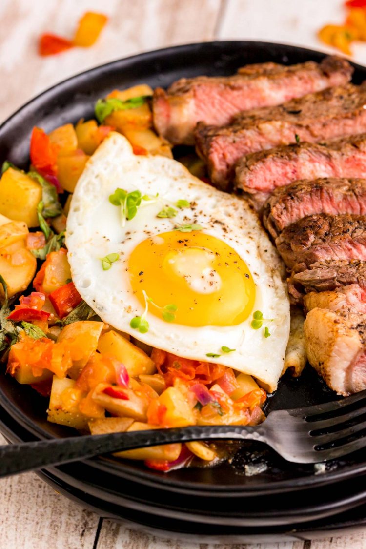 Close up photo of steak and eggs on a black plate with home fries.