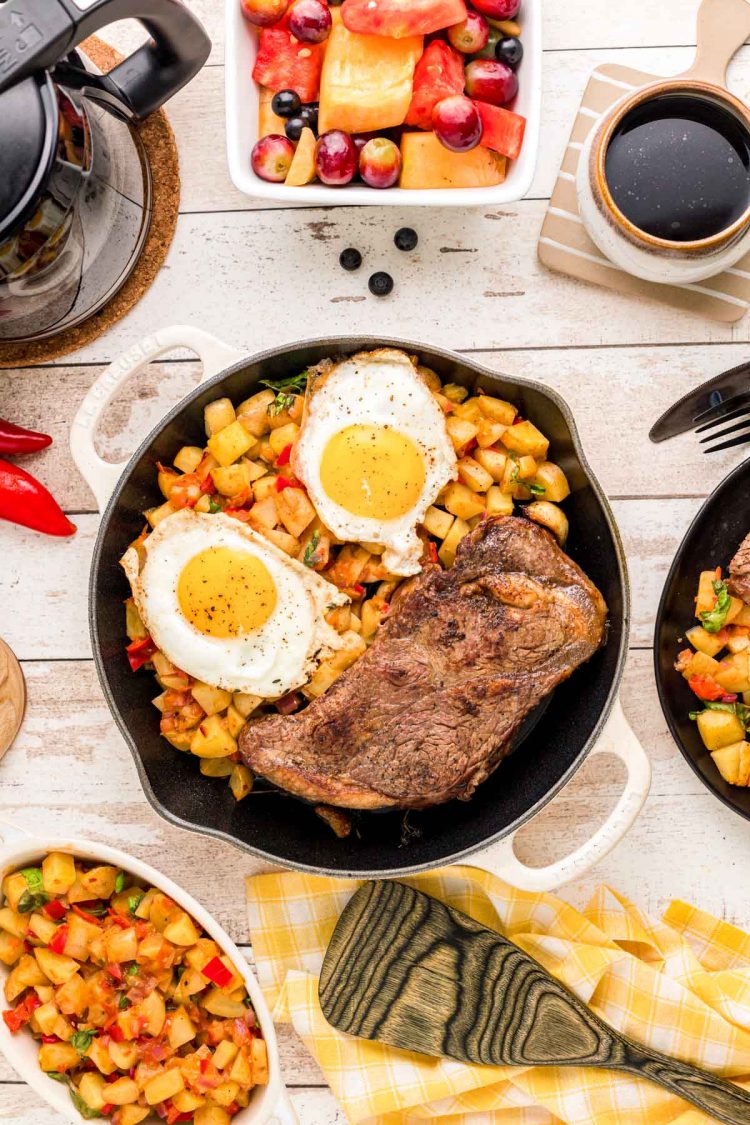 Overhead photo of steak and eggs on a skillet with home fries with coffee and plates around it.