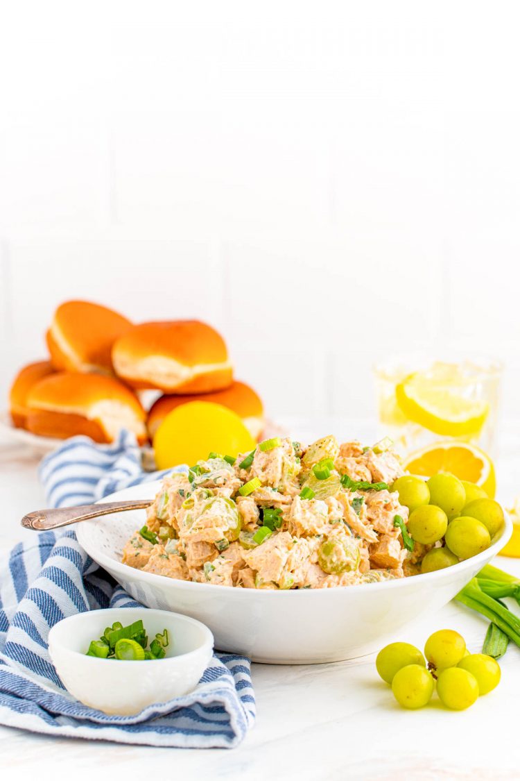 Chicken salad in a white bowl with a blue napkin under it and ingredients around the bowl on the counter.