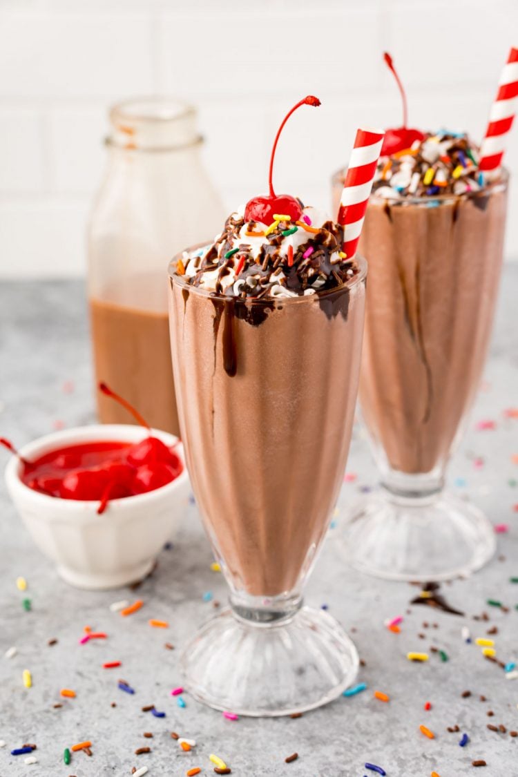 Close up photo of two milkshake glasses on a grey counter with cherries, sprinkles, and chocolate milk to the side.