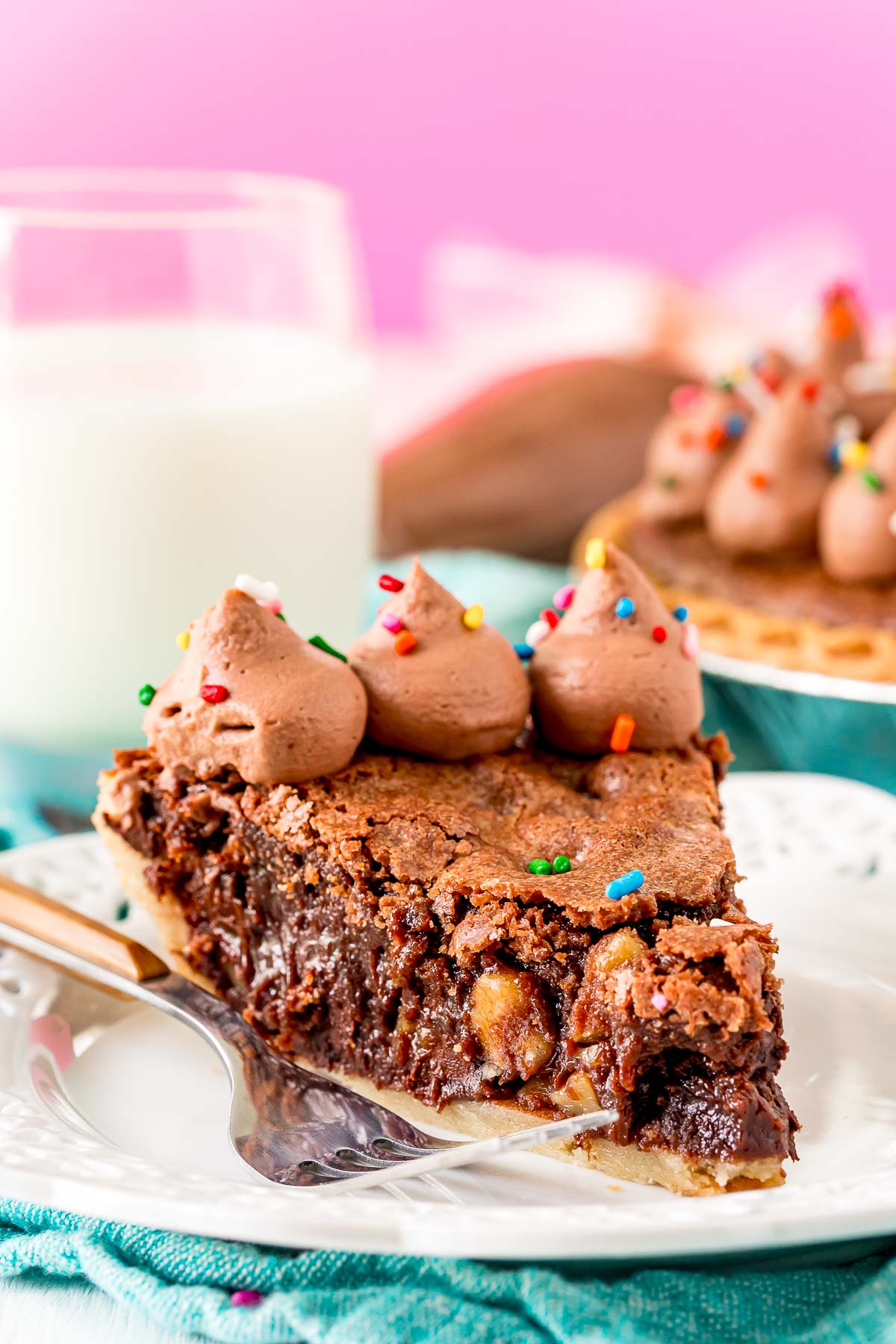 Close up photo of a slice of chocolate toll house pie on a white plate on a teal napkin with a fork next to it.