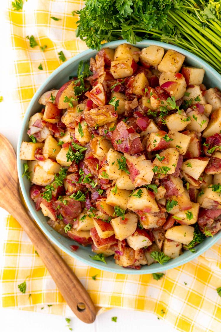 Overhead photo of German potato salad in a teal bowl on a yellow checkered napkin.