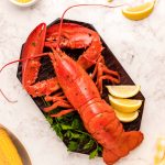 Overhead photo of a cooked lobster on a cutting board with lemon wedges and butter around it.
