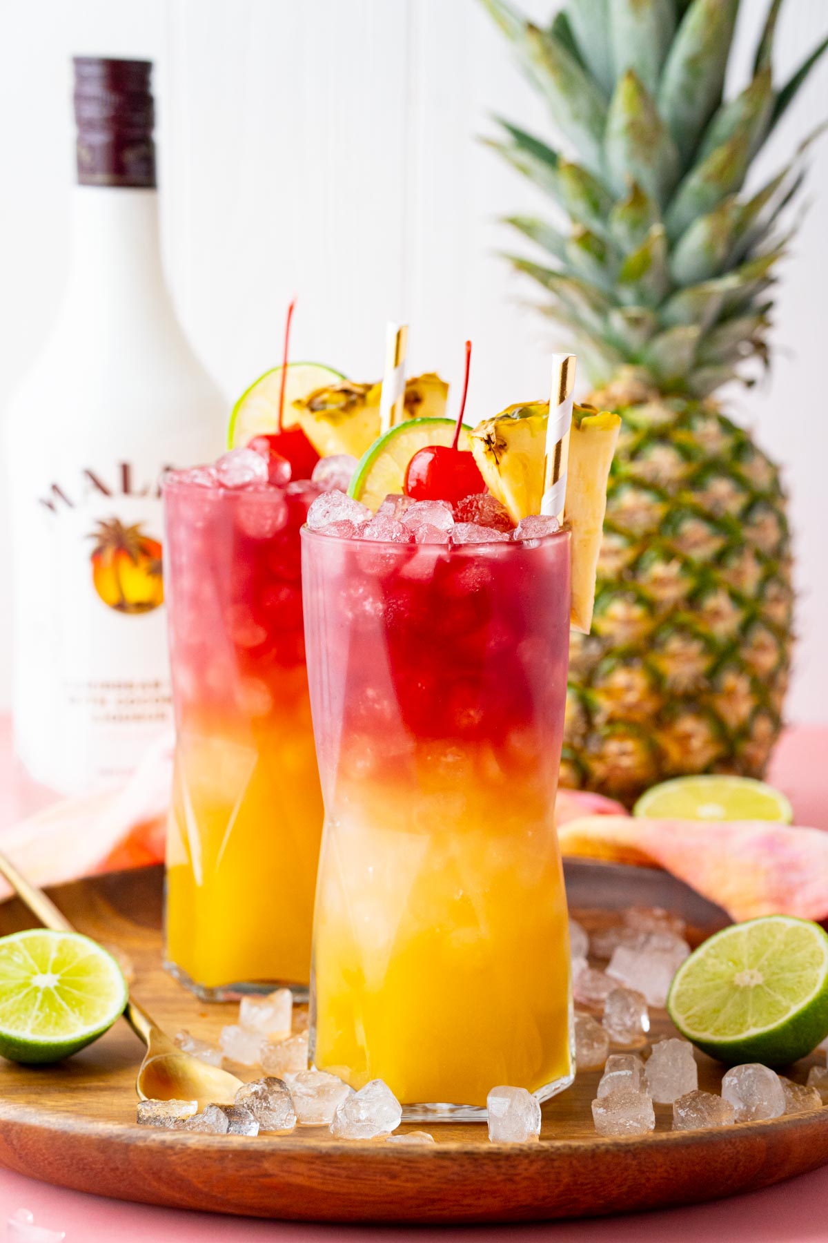 Straight on photo of two malibu bay breeze cocktails on a wooden tray with a pineapple and bottle of malibu in the background.