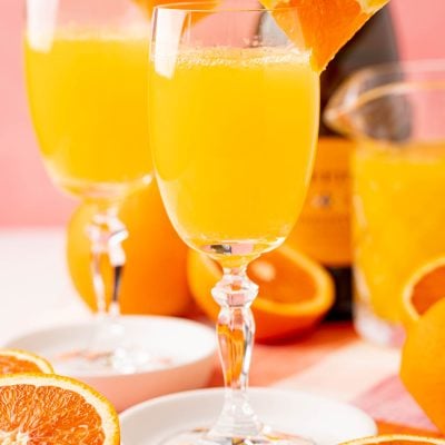 Close up photo of two mimosas on white coasters with sliced oranges around them.