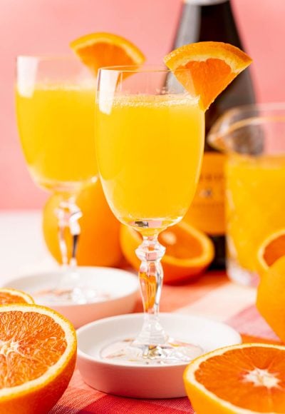 Close up photo of two mimosas on white coasters with sliced oranges around them.