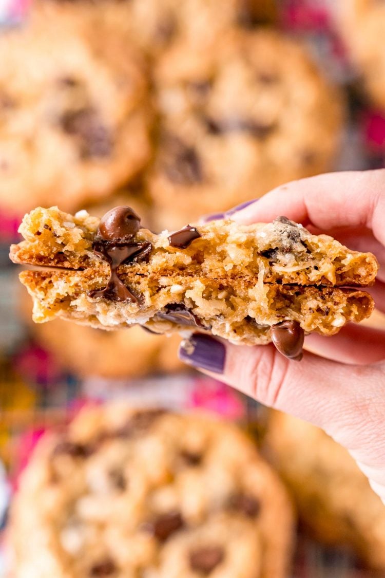 A woman's hand holding a broken in half ranger cookies to the camera so you can see the texture of the inside of the cookie.