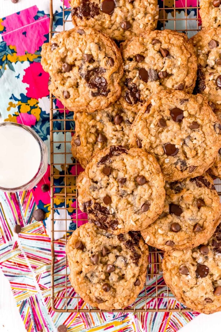 Overhead photo of ranger cookies on a wire rack on a colorful napkin with a glass of milk next to it.