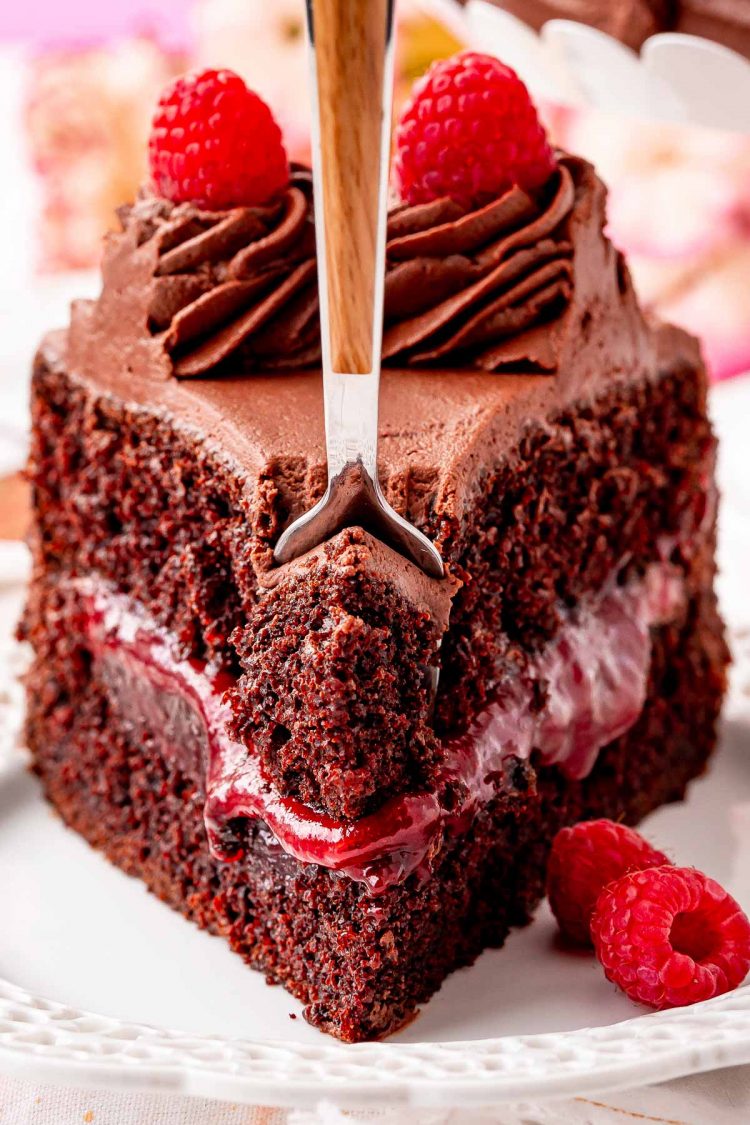 Close up photo of a fork taking a bite out of a slice of chocolate raspberry cake on a white plate.