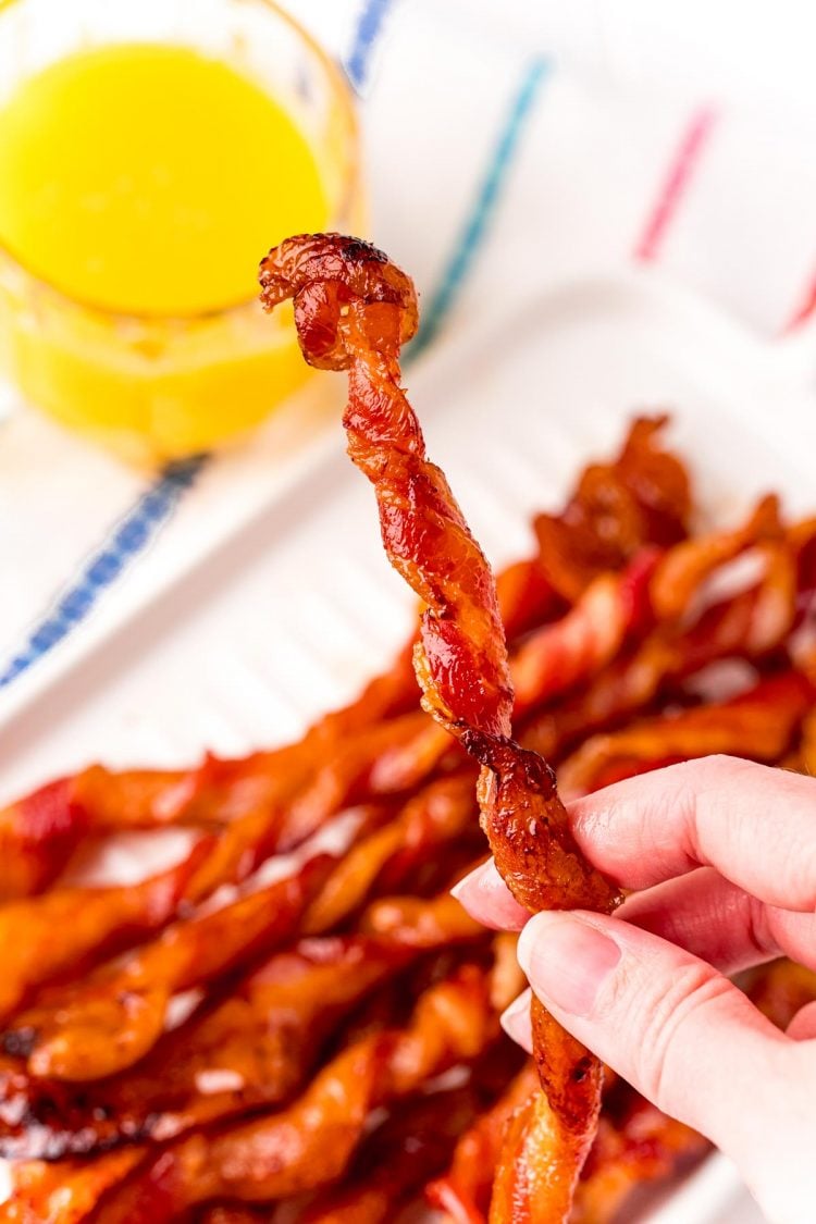 A woman's hand holding a piece of twisted bacon with more bacon and a glass of orange juice in the background.