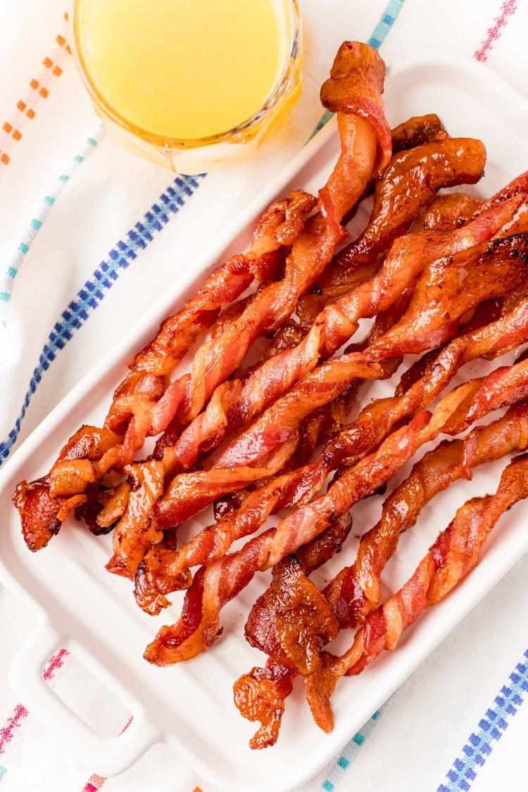 Close up photo of twisted bacon on a white plate with a glass of orange juice next to it.