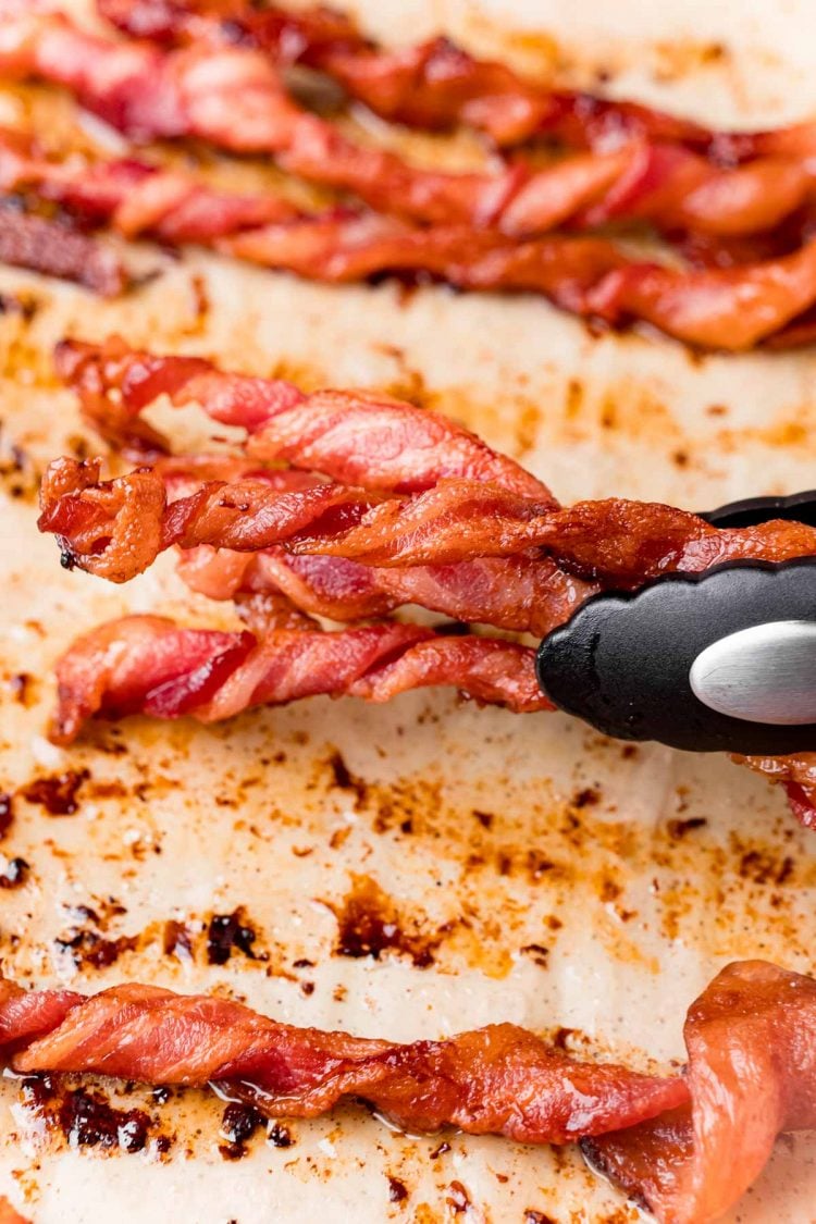 Close up photo of a pair of tongs grabbing twisted bacon off a baking sheet.