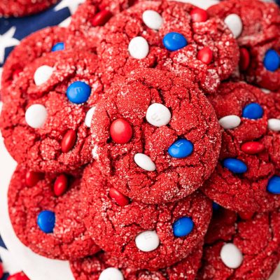 Overhead photo of red velvet cake mix cookies with red, white, and blue M&M's on a white plate on a blue star napkin.