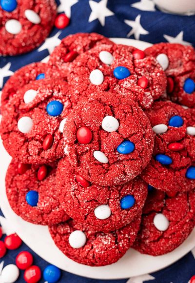 Overhead photo of red velvet cake mix cookies with red, white, and blue M&M's on a white plate on a blue star napkin.