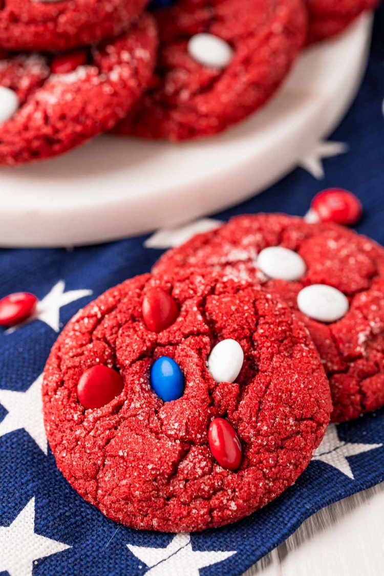 Close up photo of two red velvet cookies on a blue and white striped napkin with more cookies in the background.