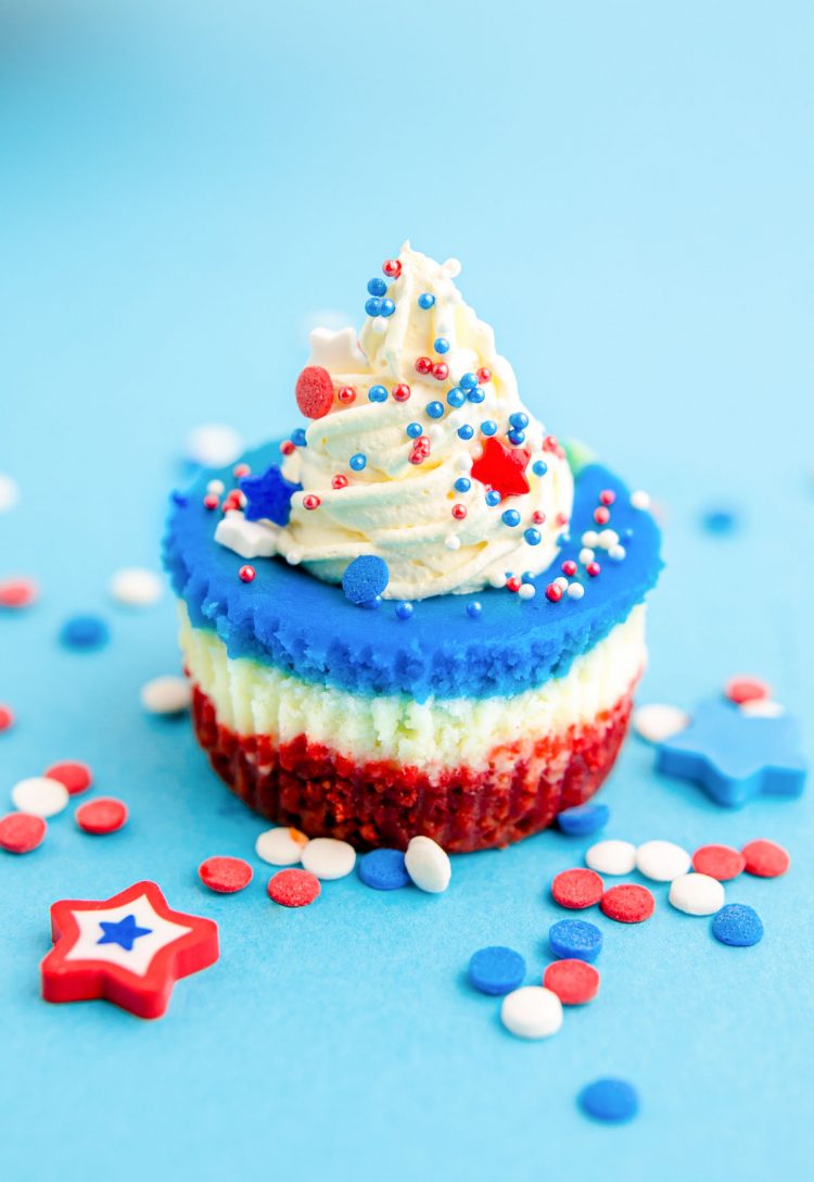 A red, white, and blue mini cheesecake sitting on a blue surface with sprinkles all around.