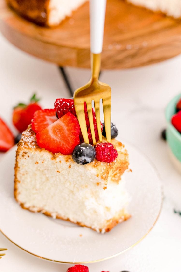 A fork taking a bite of angel food cake on a white plate topped with berries.