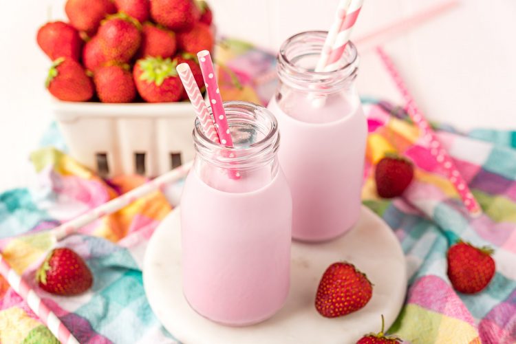 Close up horizontal photo of two milk bottles with strawberry milk and pink straws in them on a white board on a colorful napkin.