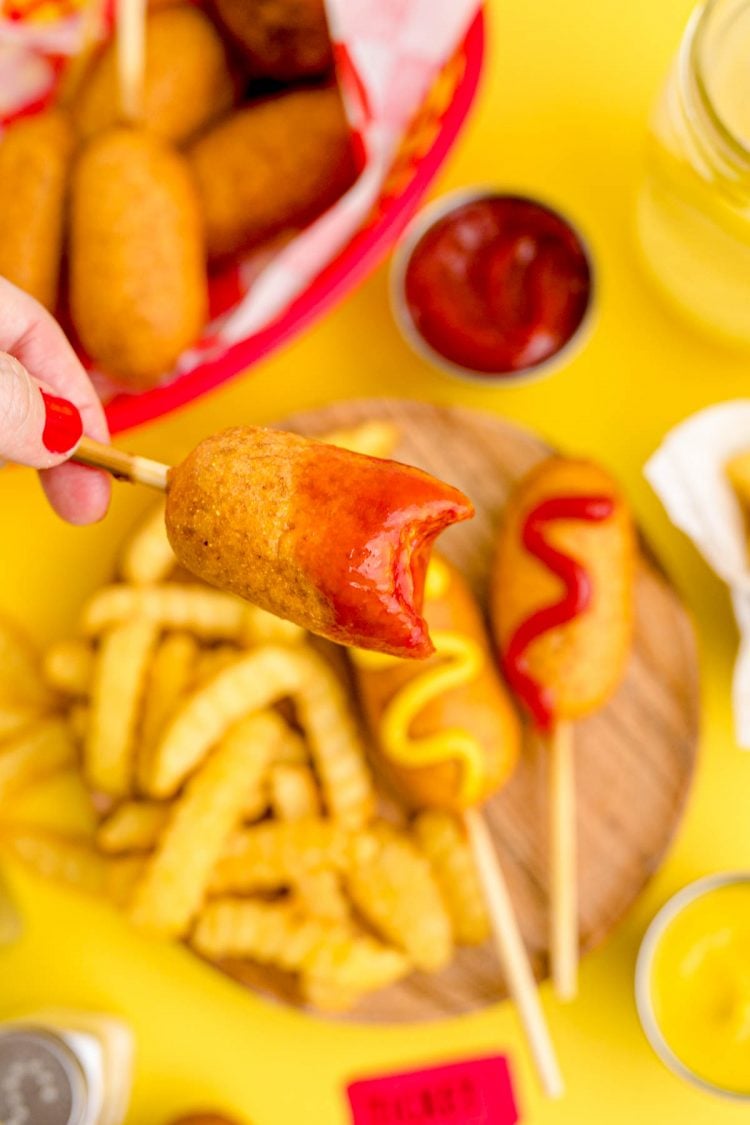 A woman's hand holding a mini corn dog with a bite missing close to the camera.