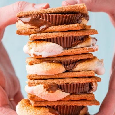 Close up photo of a stack of s'mores being held by a woman's hand in front of a blue background.