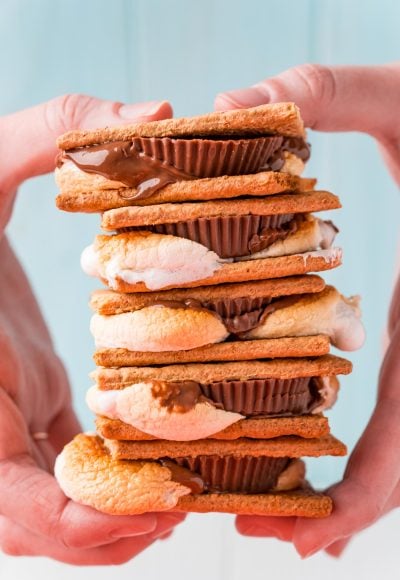 Close up photo of a stack of s'mores being held by a woman's hand in front of a blue background.