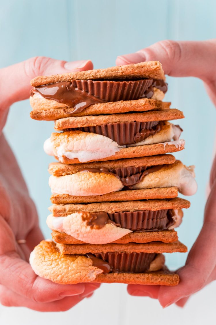 Close up photo of a stack of s'mores being held by a woman's hand in front of a blue background.