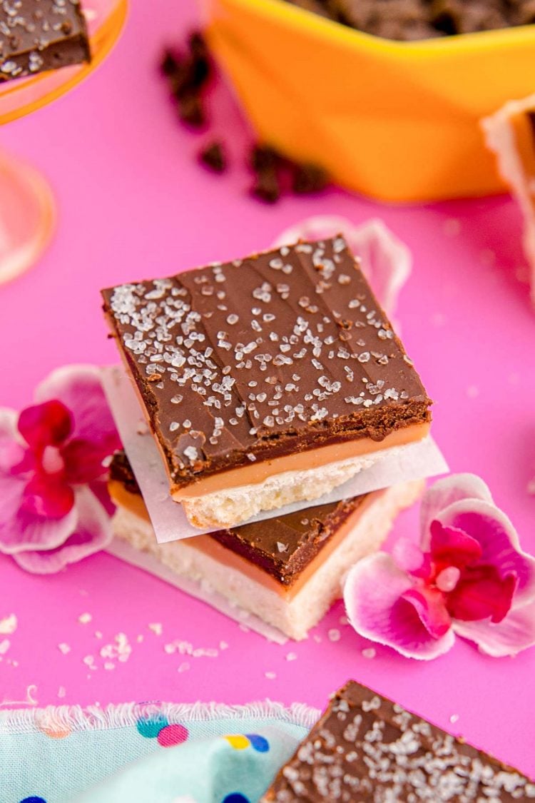 Two Millionaire shortbread bars stacked on each other on a pink surface with flowers around them.